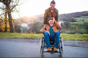 A young man and his senior father in wheelchair on a walk in town at sunset.