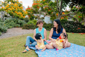 Grandmother, daughter, and two granddaughters on blue quilt on lawn with beautiful landscaping in background.