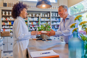 Photo of positive woman druggist wearing white coat giving advice to customer in pharmacy.  Medicine, pharmaceutics, health care and people concept - Apothecary and senior man customer buying drug at drugstore. Medicine, pharmaceutics, health care and